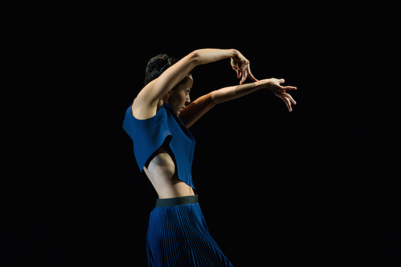 A female dancer in profile extends her left arm at eye level while her right arm bends at the elbow over her head. Her right fingers splay and rest on her left forearm